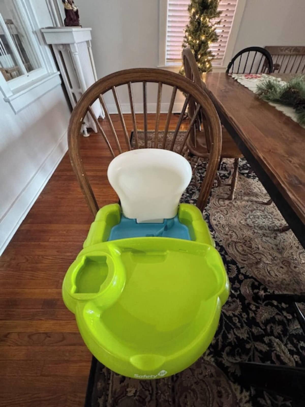 A dining area with a wooden dining table, a green and blue high chair, and a Christmas tree in the background.