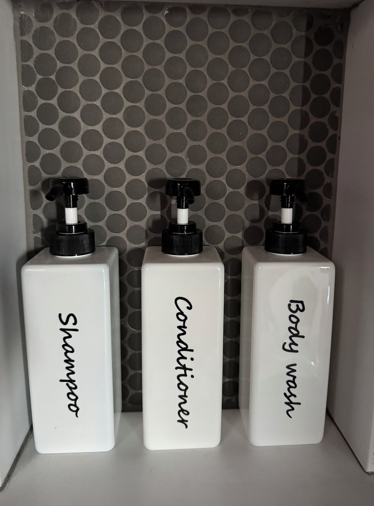 Three white bottles labeled 'Shampoo,' 'Conditioner,' and 'Body wash' in a white cabinet with a polka dot pattern.