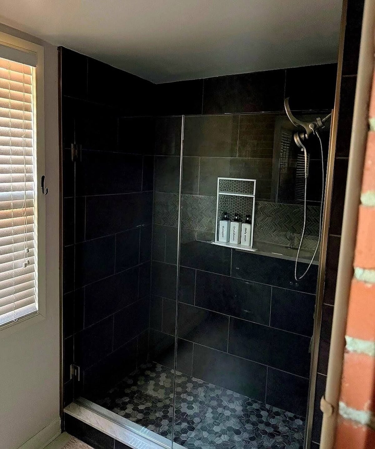 A glass-enclosed shower with dark tiles and a small shelf with toiletries.