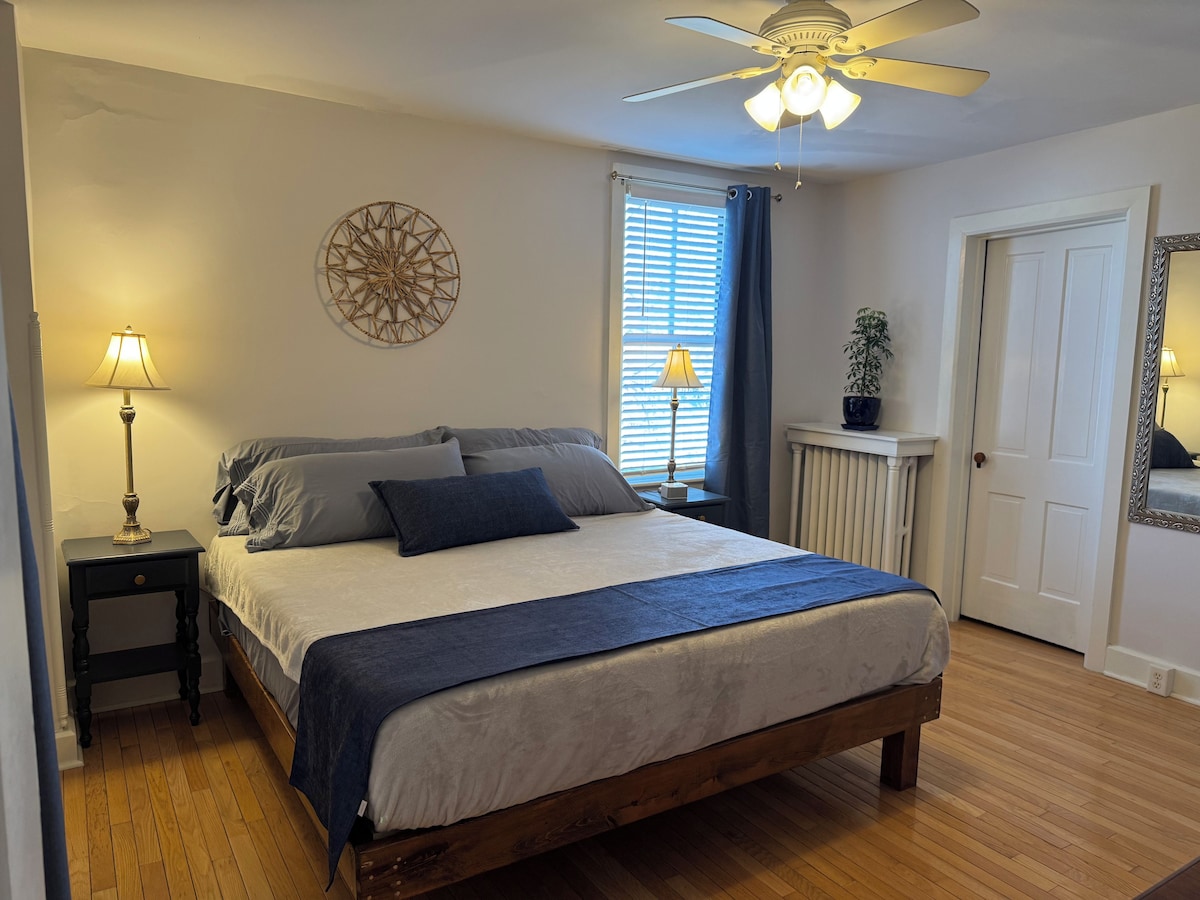 A bedroom with a bed, a nightstand, a ceiling fan, a window with blinds, and a door.