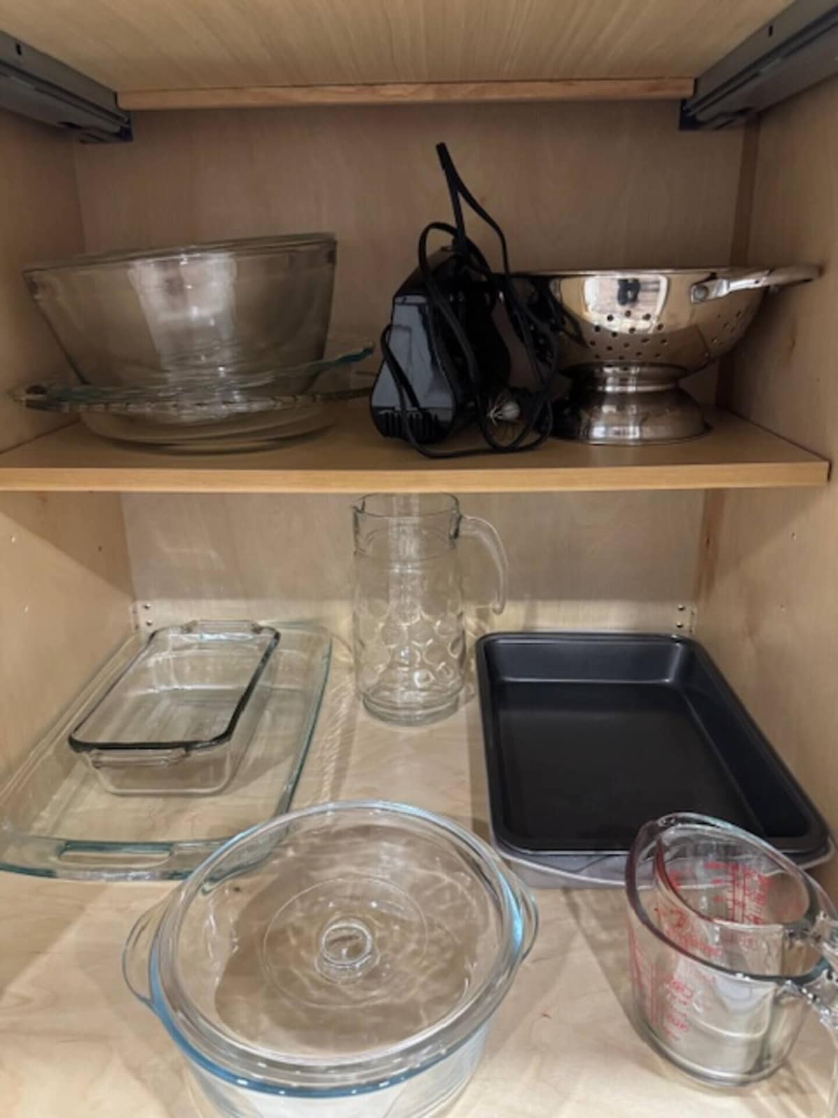 A kitchen shelf with various glassware and cookware, including a glass measuring cup, a glass bowl, and a metal colander.