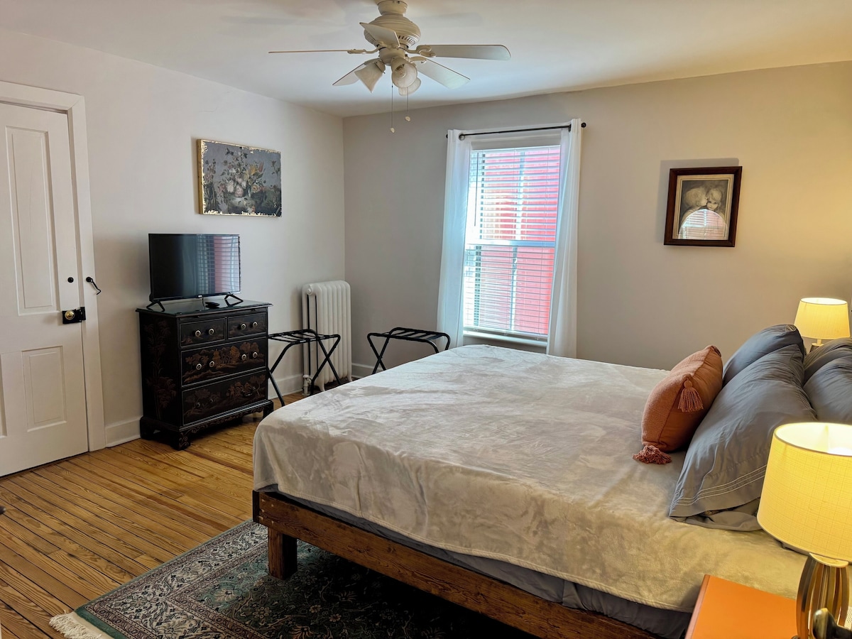 A bedroom with a bed, a dresser, a television, a radiator, a ceiling fan, and a window with blinds.