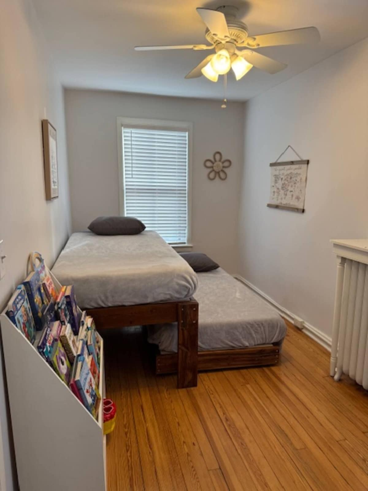 A small, cozy bedroom with a single bed, a nightstand, a bookshelf, a ceiling fan, and a window with blinds.