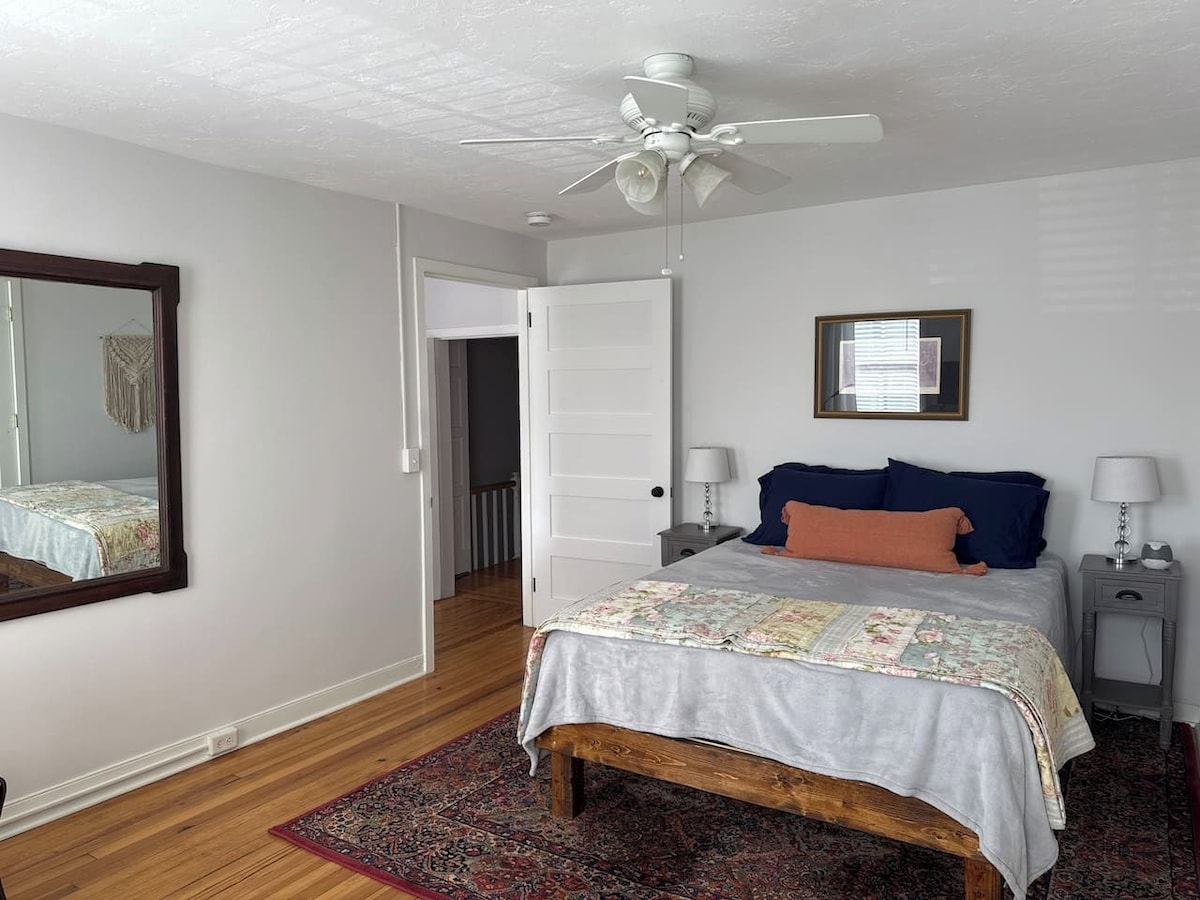 A bedroom with a bed, a ceiling fan, a nightstand, a bedside table, a mirror, and a door leading to another room.