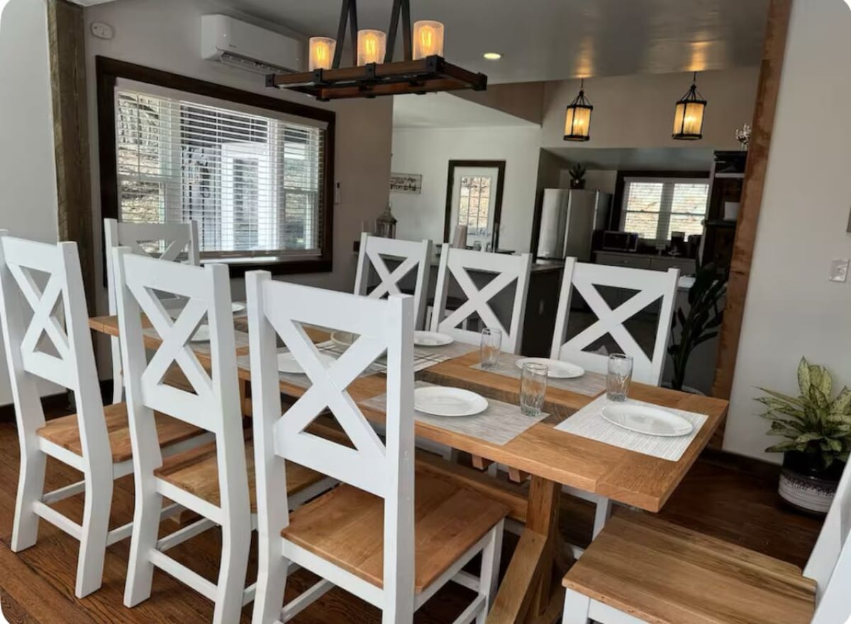A dining area with white chairs and a wooden table, a chandelier, and a view of the kitchen in the background.