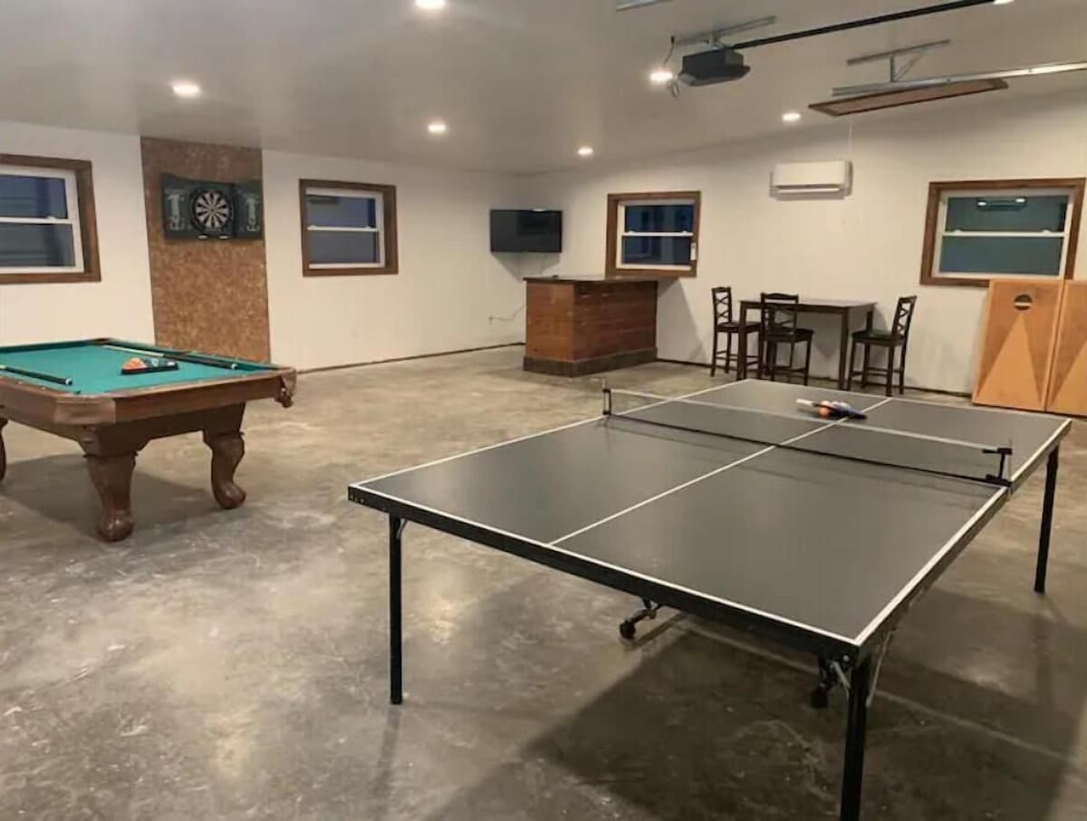 A room with a pool table, ping pong table, dartboard, and a small bar area with a TV.