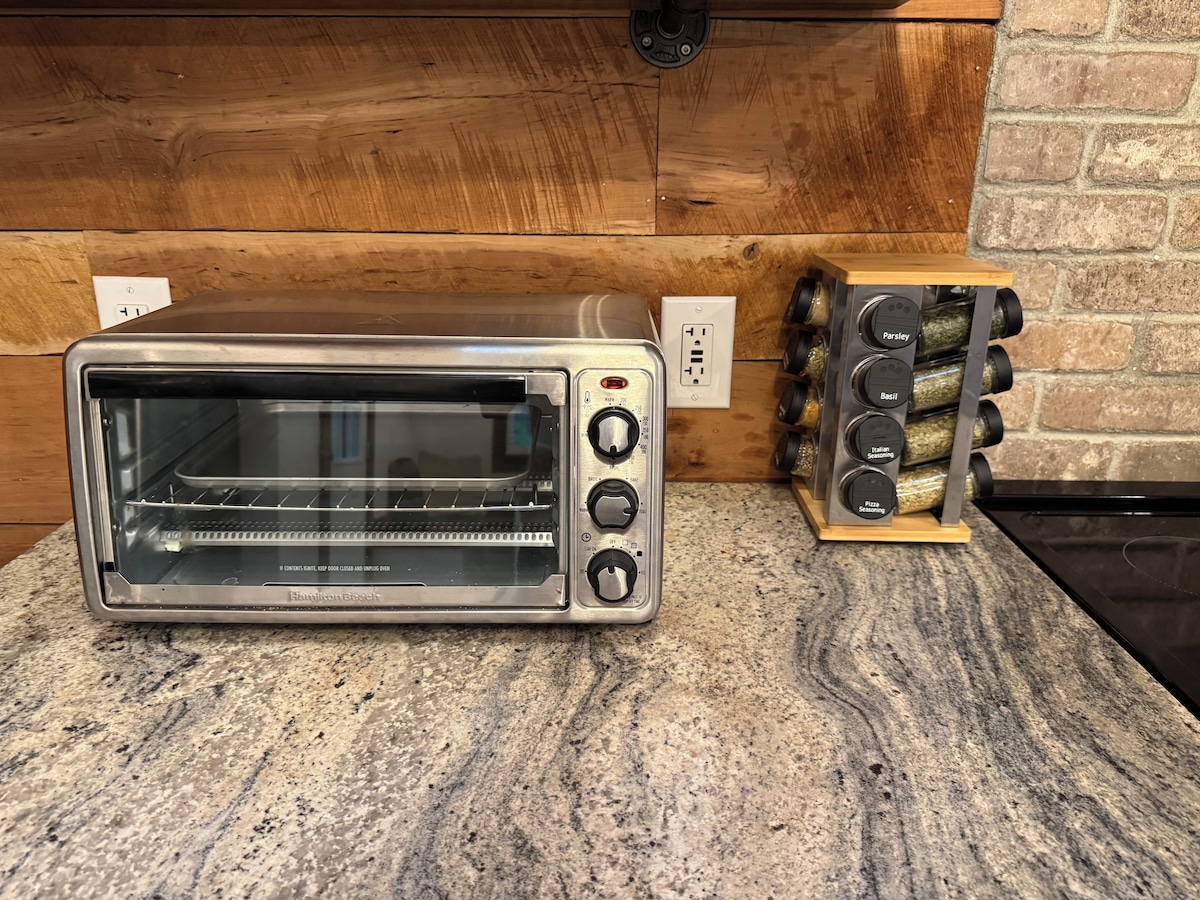 A Hamilton Beach toaster oven on a granite countertop with a spice rack and a wooden wall in the background.