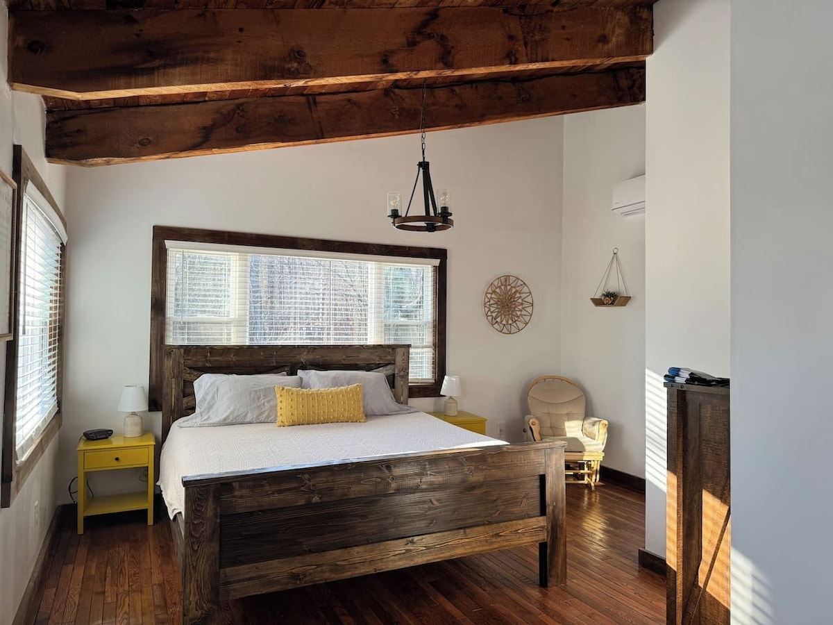 A bedroom with a wooden bed, a nightstand, a chair, a window with blinds, and a wall-mounted light fixture.