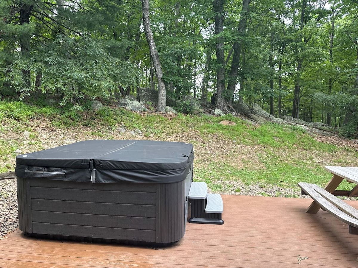 A hot tub with a cover, a bench, and a picnic table in a wooded outdoor setting.